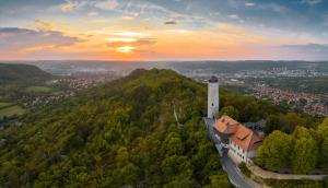 Blick aus der Vogelperspektive auf einen historischen Bergfried, der sich auf einem Berg befindet. Im dahinter gelegenen Tal erstreckt sich die Stadt Jena im Abendlicht.
