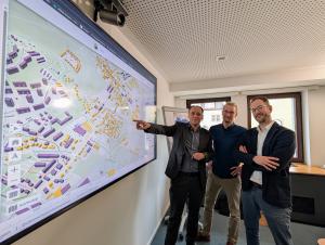 Drei Männer stehen in einem Büroraum vor einem großflächigem Wandbildschirm, auf dem der kommunale Wärmeplan zu sehen ist.