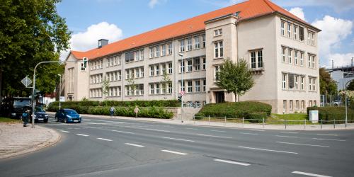 Blick auf eine Kreuzung. Auf der Linkesn Seite stehen Uatos an einer Ampel. Im Hintergund ist ein großes Gebäude mit einem roten Spizdach zu sehen.
