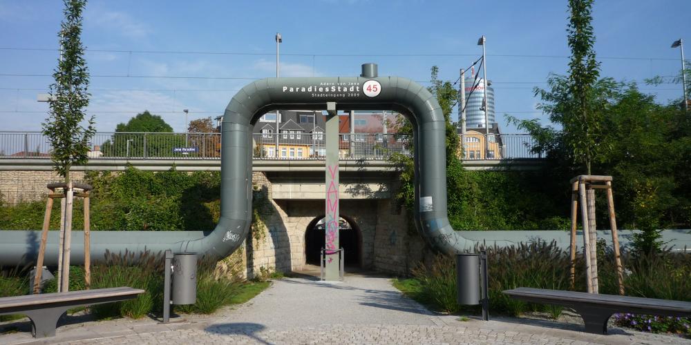 Eine Fernwärmeleitung verläuft ebenerdig und über den Eingang einer Unterführung über einen Bahnschiene. Im Hintergund ist der Jentower zu sehen. f