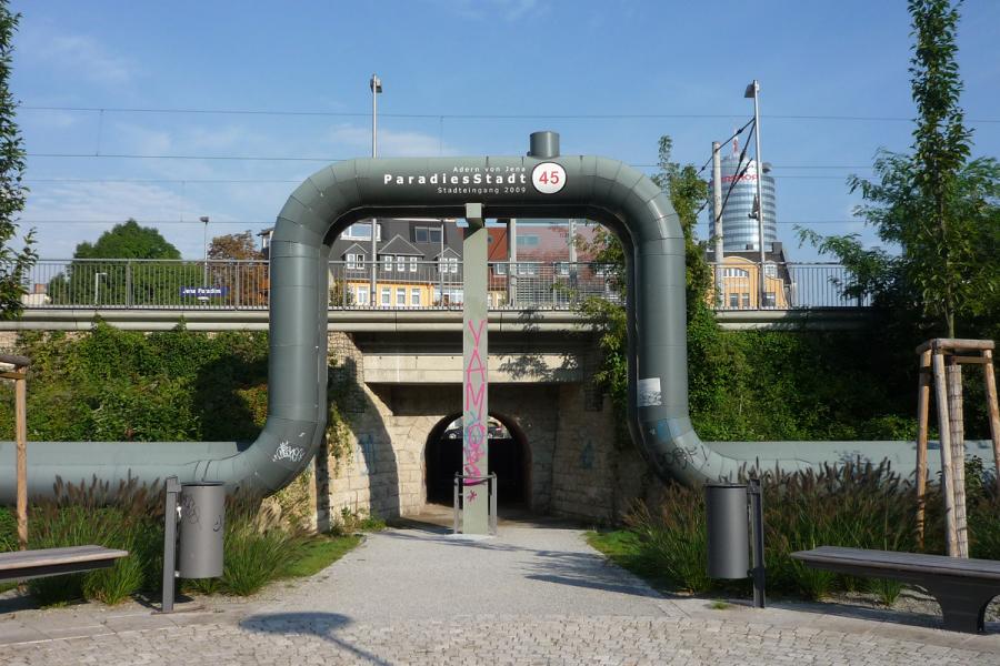 Eine Fernwärmeleitung verläuft ebenerdig und über den Eingang einer Unterführung über einen Bahnschiene. Im Hintergund ist der Jentower zu sehen. f