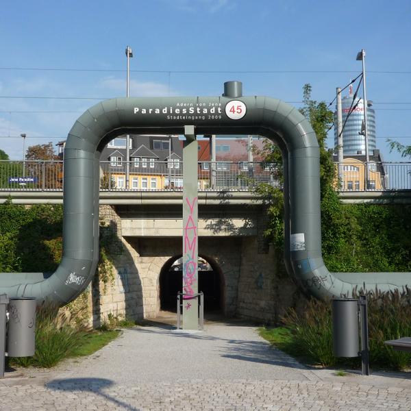 Eine Fernwärmeleitung verläuft ebenerdig und über den Eingang einer Unterführung über einen Bahnschiene. Im Hintergund ist der Jentower zu sehen. f
